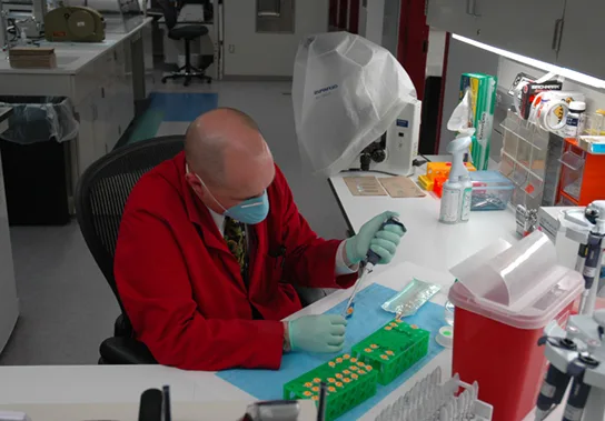 A forensic scientist in a lab coat, mask, and gloves works at a bench lined with sample tubes and lab equipment.