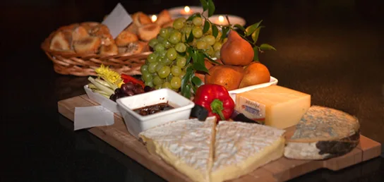 A wooden board holding wedges of several cheeses, a red bell pepper, a small dish of dark preserve, green grapes, and pears, with a basket of bread rolls behind.