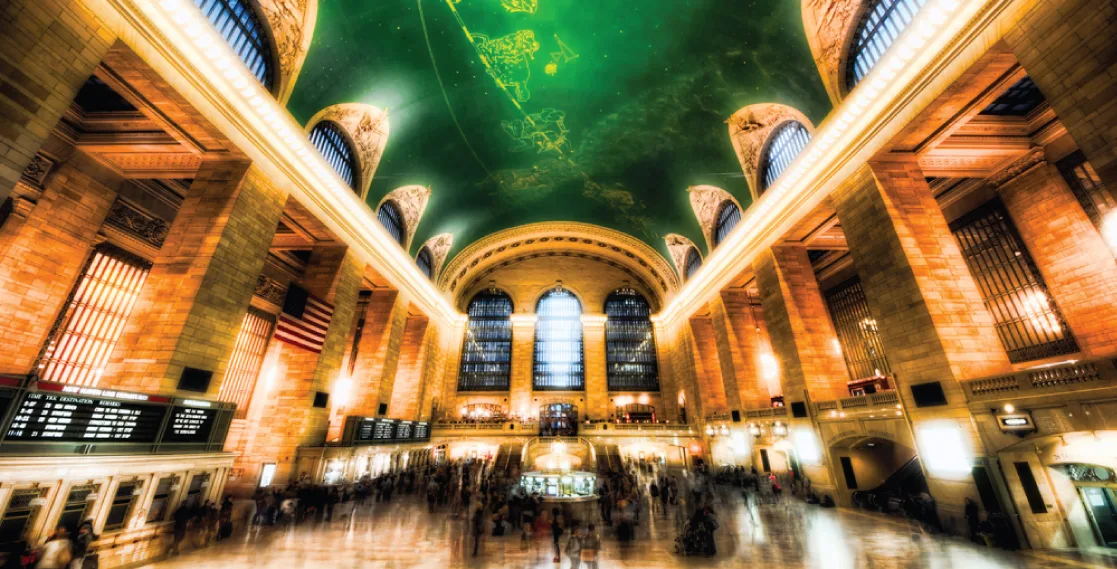 The ornate marble concourse of Grand Central Terminal, its tall arched windows and gilded moldings lit gold, a green ceiling painted with constellations overhead, and crowds of people moving across the floor below.