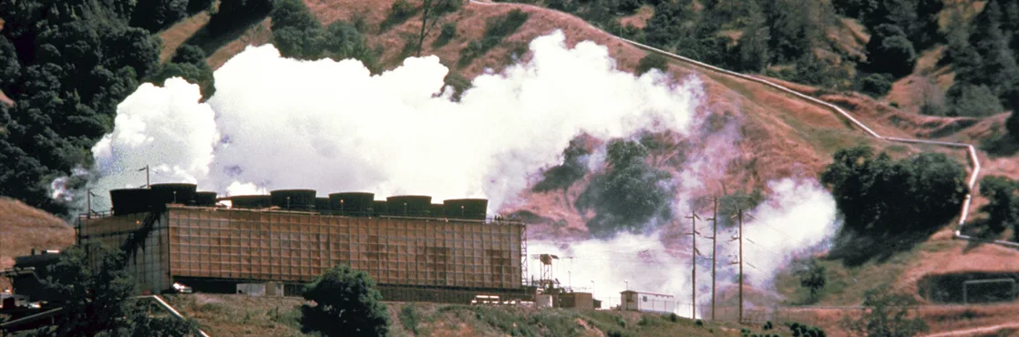 A geothermal power plant's long building sits on a green, tree-covered hillside, with thick white steam billowing from cooling towers on its roof and drifting across the slope behind it.