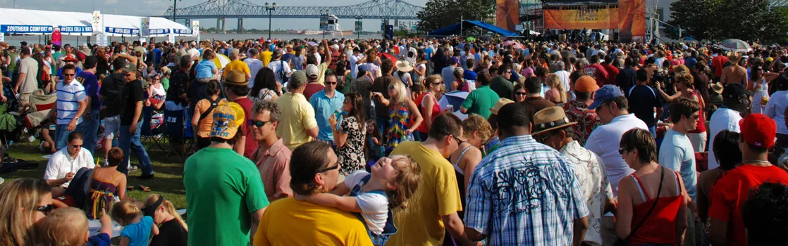 A dense, sunlit outdoor crowd fills a riverside festival ground, with a stage and tents in the background and a bridge over water visible beyond them.