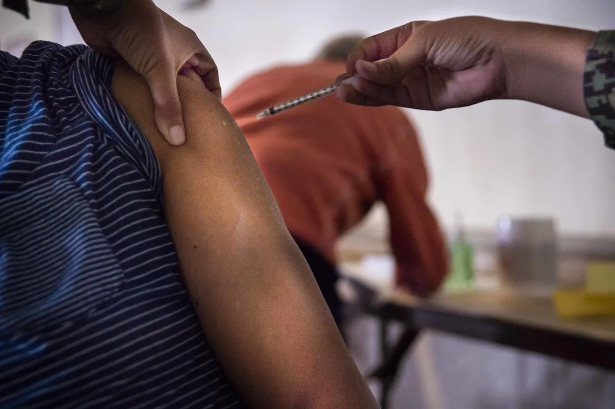 A close-up photo of a person's upper arm as another person's hand, wearing a camouflage-patterned sleeve, gives them an injection with a syringe.