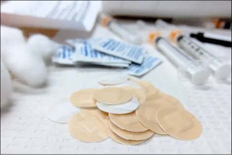 A close-up photograph of medical supplies on a table: several capped syringes, a stack of individually wrapped alcohol swabs, cotton balls, and a pile of round adhesive bandages in the foreground.