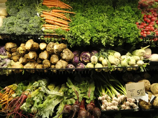 A grocery produce display with kale, carrots, and radishes on the top shelf, kohlrabi bulbs and turnips on a middle shelf, and beets, garlic, and leafy greens on the bottom shelf.