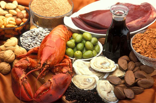 A close-up photo of a spread of foods on an orange cloth: a bowl of mixed nuts and whole walnuts at the upper left, a bowl of pale granular wheat germ, a dish of sunflower seeds, a whole cooked red lobster, a bowl of green olives, opened bivalve shells, a glass bottle of dark liquid, slices of dark-red raw liver on a plate, a bowl of ground cocoa at the right, a pile of Brazil nuts at the lower right, and scattered black peppercorns in front.