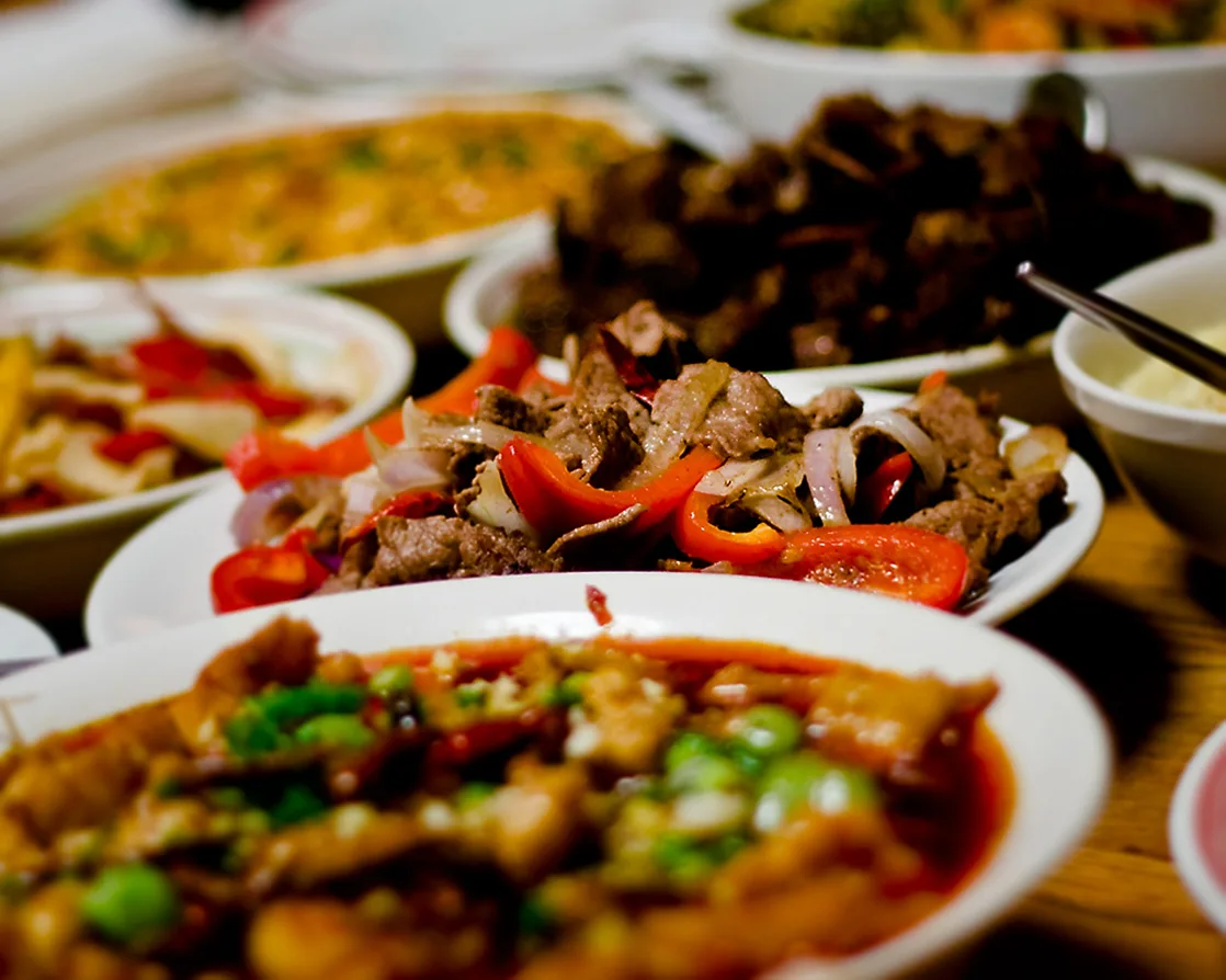 Several white bowls and plates crowd a wood table, each holding a different stir-fried or saucy dish — sliced beef with red peppers and onions in one bowl, a pea-and-meat dish in a reddish sauce nearest the camera, and blurred dishes further back.