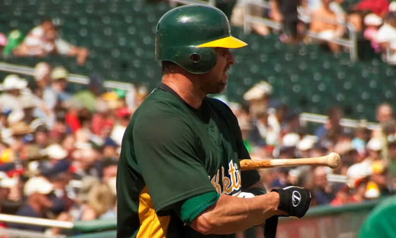 Baseball player Jason Giambi, wearing a green batting helmet and an Oakland Athletics jersey, holds a bat in the batter's box with a crowd filling the stands behind him.