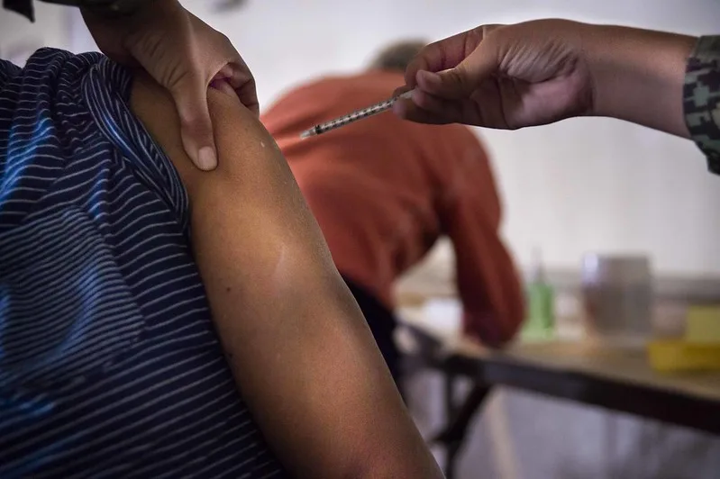 A close-up photo of a person's upper arm being injected with a syringe held by a gloved hand, with another person seated in the background.