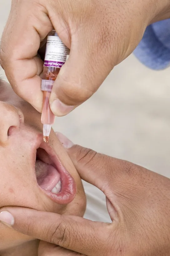 A close-up photo of a person administering oral vaccine drops from a small dropper bottle into an infant's open mouth.