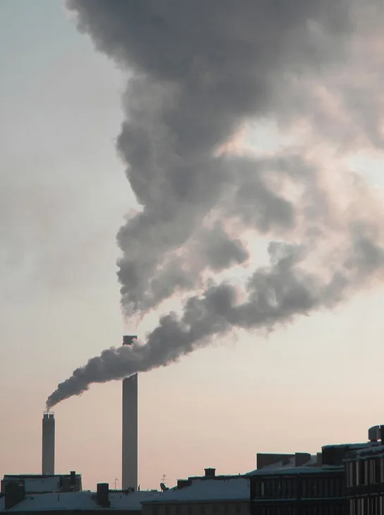 Two industrial smokestacks rise above a row of buildings, one releasing a large plume of dark gray smoke that spreads across the sky and the other releasing a smaller plume of white smoke.