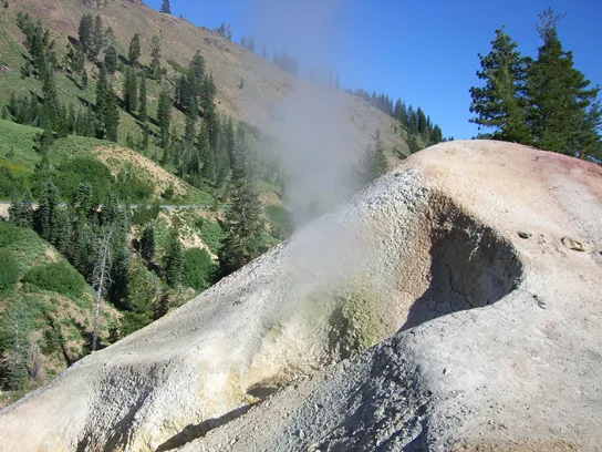 A white, pyramid-shaped mineral mound sits on a forested hillside, with gray steam escaping from an opening near its base.