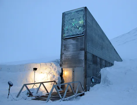 Photo of a tall, wedge-shaped concrete structure with a glowing triangular panel at its peak and a lit doorway at ground level, built into a snow-covered hillside, with floodlights and a railed walkway leading to the entrance.