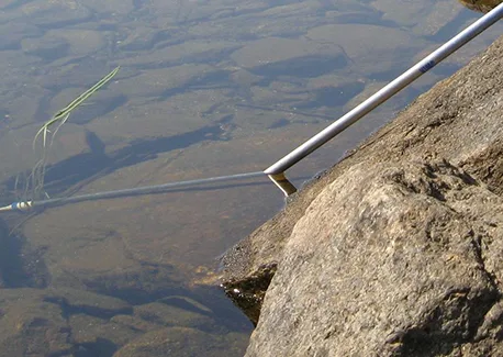 A photograph looking down at a metal pole resting against a rock at the edge of shallow water. Where the pole crosses the waterline, its submerged portion appears to angle sharply compared with the straight portion above the surface, creating the illusion that the pole bends at the water's surface.