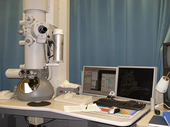 A photograph of a large transmission electron microscope: a tall, column-shaped instrument on a bench beside a desk with a keyboard, mouse, and two monitors displaying diagnostic screens.