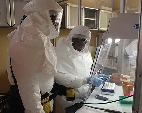 A photograph of two researchers fully covered in white biohazard suits with hoods and face coverings, working with laboratory samples inside a ventilated hood.