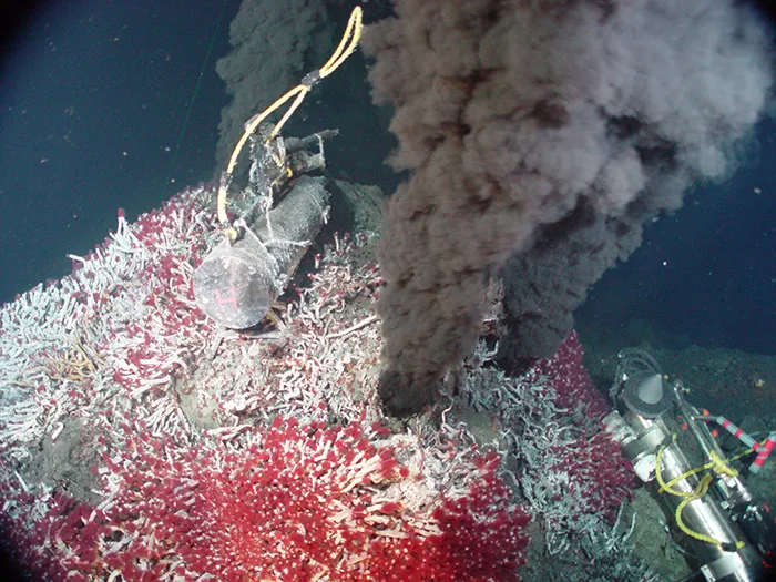 An underwater photo of a hydrothermal vent chimney billowing a dark, mineral-laden plume from its top, with clusters of red tubeworms and pale organisms encrusting the rock around its base and scientific sampling instruments positioned nearby.