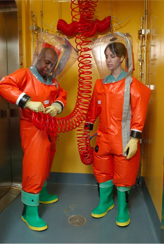 A photo of two lab workers in orange positive-pressure suits with clear plastic hoods over their heads, connected by a shared red coiled air hose, standing on a grated floor in a yellow-walled decontamination area.
