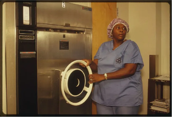 A color photograph of a woman in blue scrubs and a cloth cap, holding open the round hinged door of a large stainless-steel autoclave unit in a laboratory hallway.