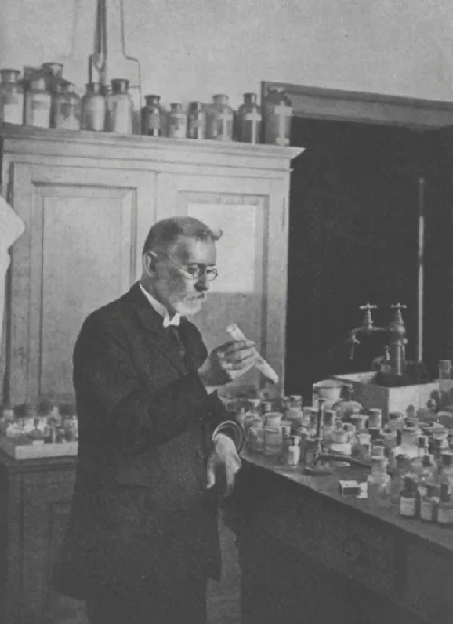 A black-and-white photograph of an older, bearded man in a dark suit standing in a laboratory, holding a small vial up in one hand. Behind him is a tall cabinet topped with a row of glass bottles, and a counter to his right holds many more small bottles beside a sink with two taps.