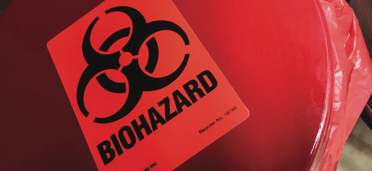 A close-up of a red biohazard waste bin with a rectangular orange-red warning label showing the black biohazard trefoil symbol above the word BIOHAZARD, and a red plastic liner bag visible curling over the bin's rim.