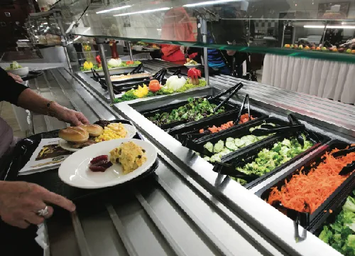 Photo of a cafeteria food line with a glass sneeze guard mounted above open trays of salad and other prepared food.